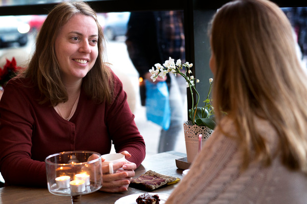 En ung kvinna sitter mitt emot en annan ung kvinna vid ett bord på ett kafé. Hon ler. I handen håller hon en mugg och mellan kvinnorna står ett fat med en kaka.
