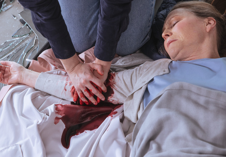 En kvinna med kraftigt sår på ena armen ligger på golvet medan två händer trycker på blödningen.